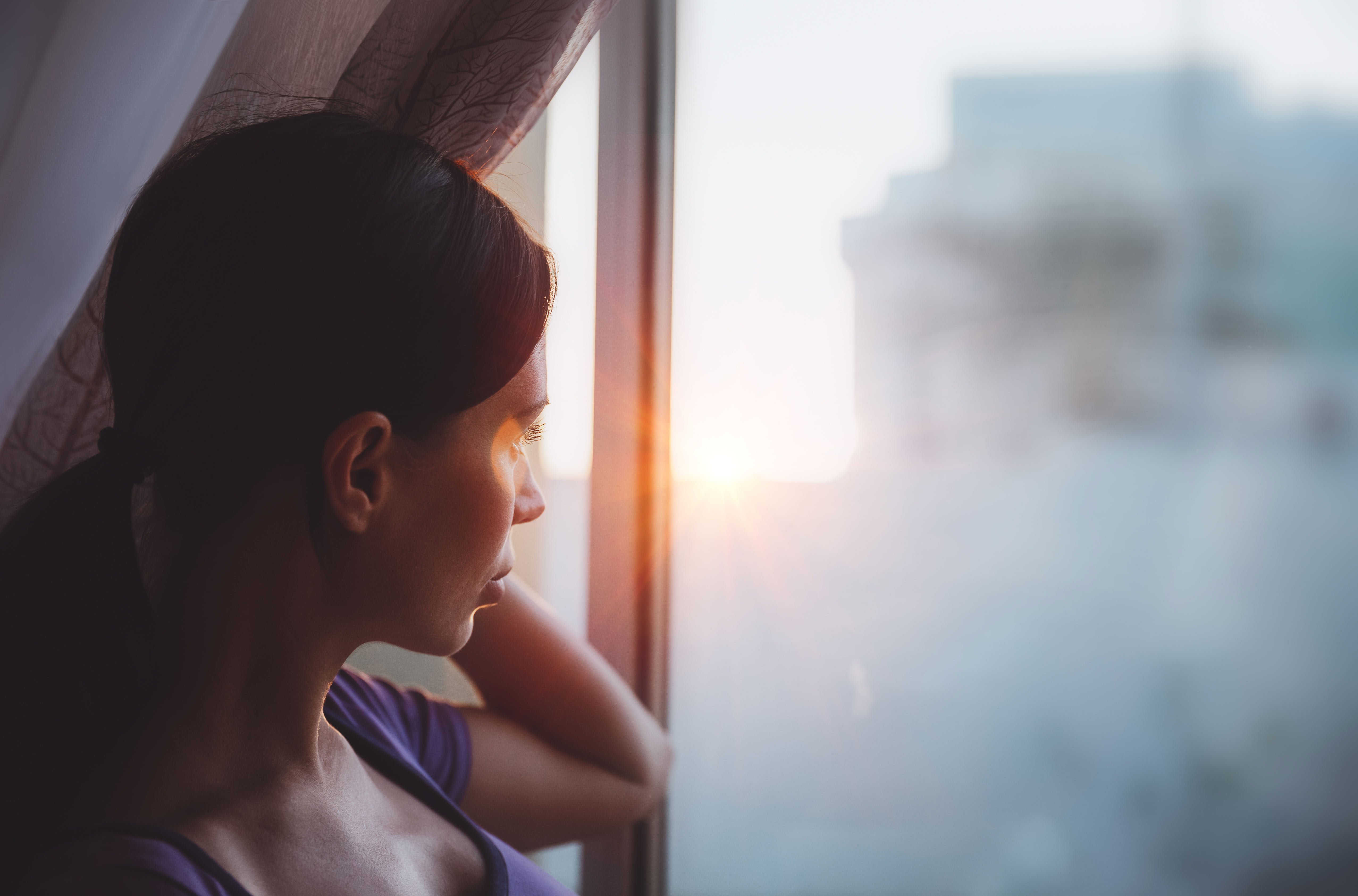 477082000079016003 zc v1 1790314350360 woman gazing out window at sunrise in city 2026 09 23 20 39 00 utc (1)